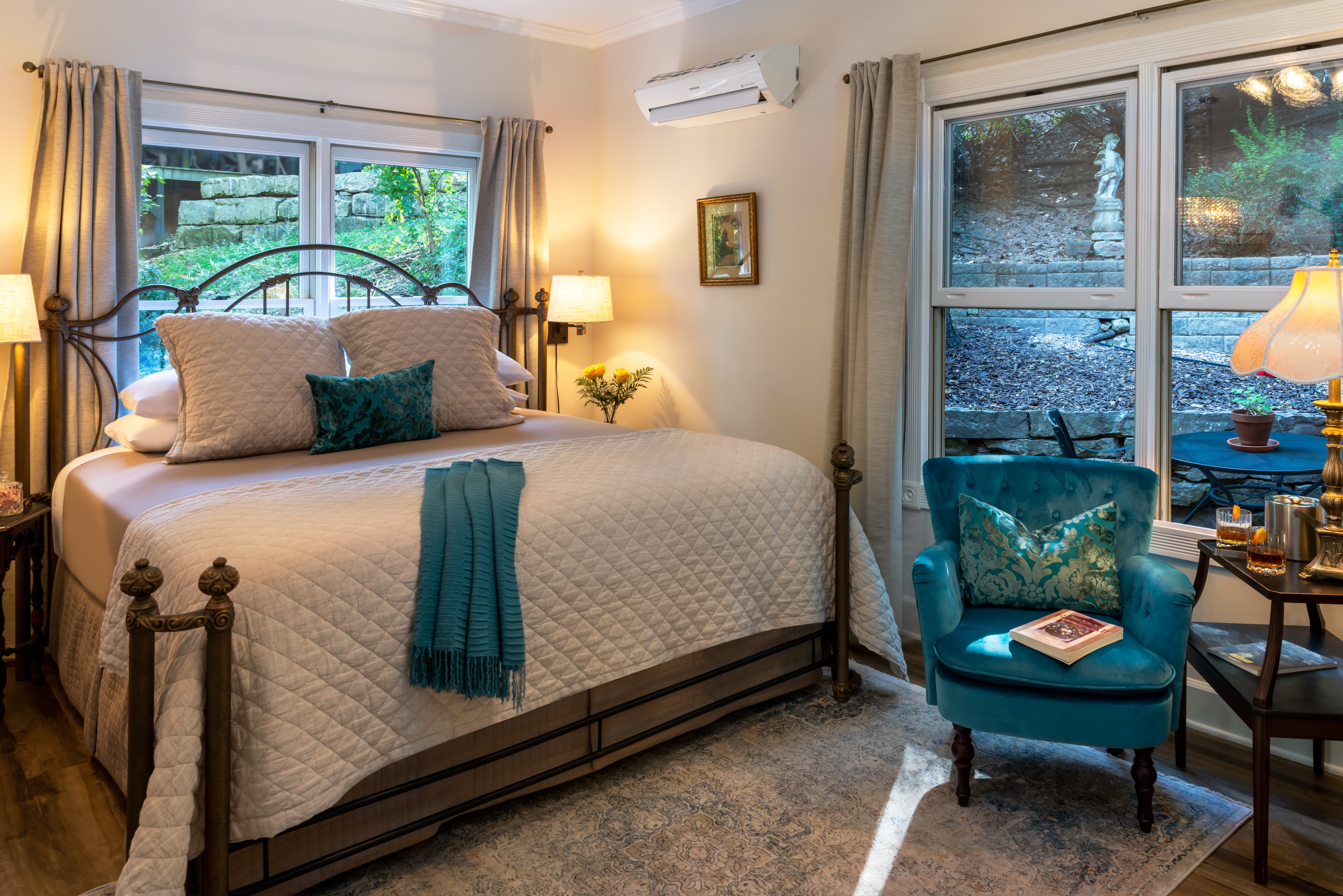 A cozy bedroom featuring a large bed, blue accent chair, and natural light from windows.