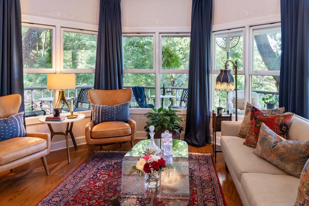 A cozy living room featuring large windows with a view of nature, two armchairs, a white sofa, and a decorative coffee table.