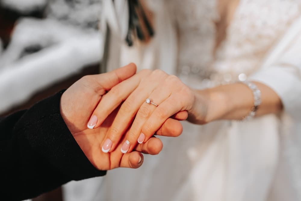 A close-up of two hands holding each other, one adorned with a simple engagement ring.
