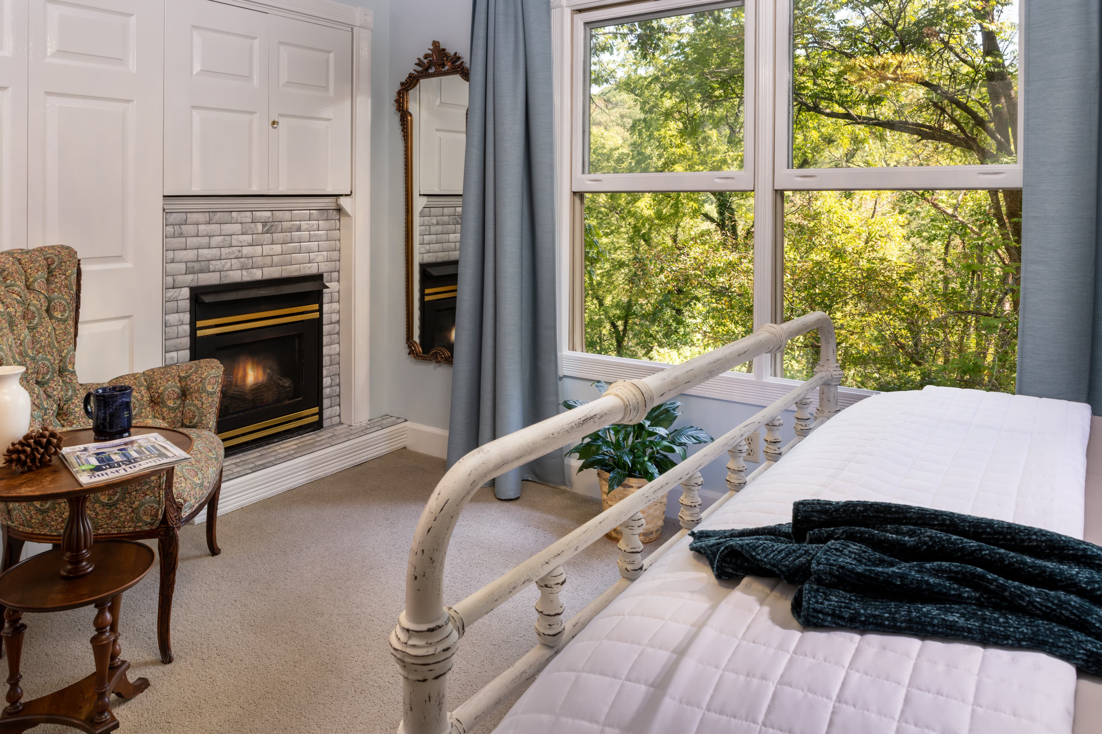 A cozy bedroom featuring a bed, a fireplace, a mirror, and large windows overlooking greenery.