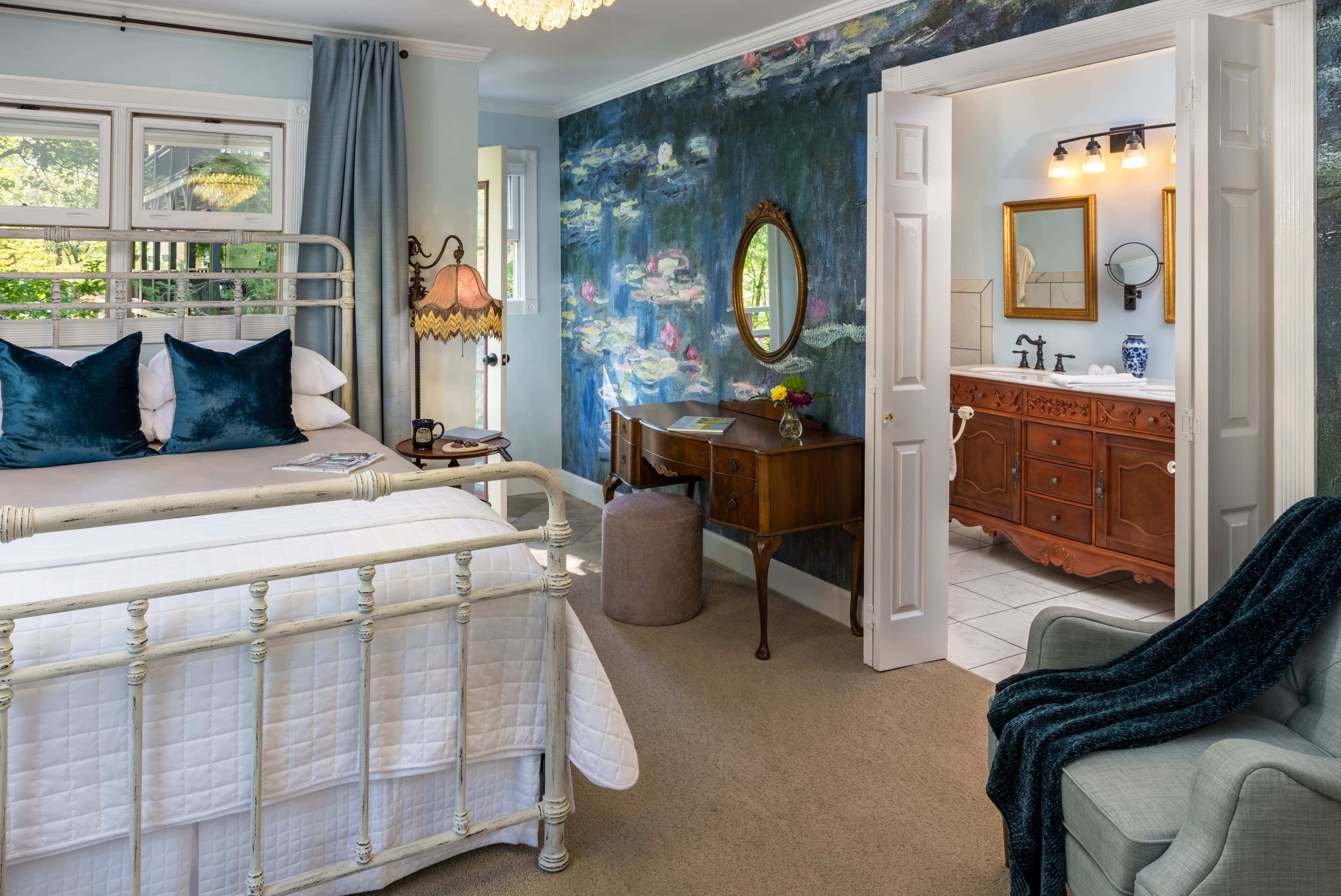 A cozy bedroom featuring a vintage metal bed, elegant decor, and a colorful mural, with an adjoining bathroom visible through an open door.