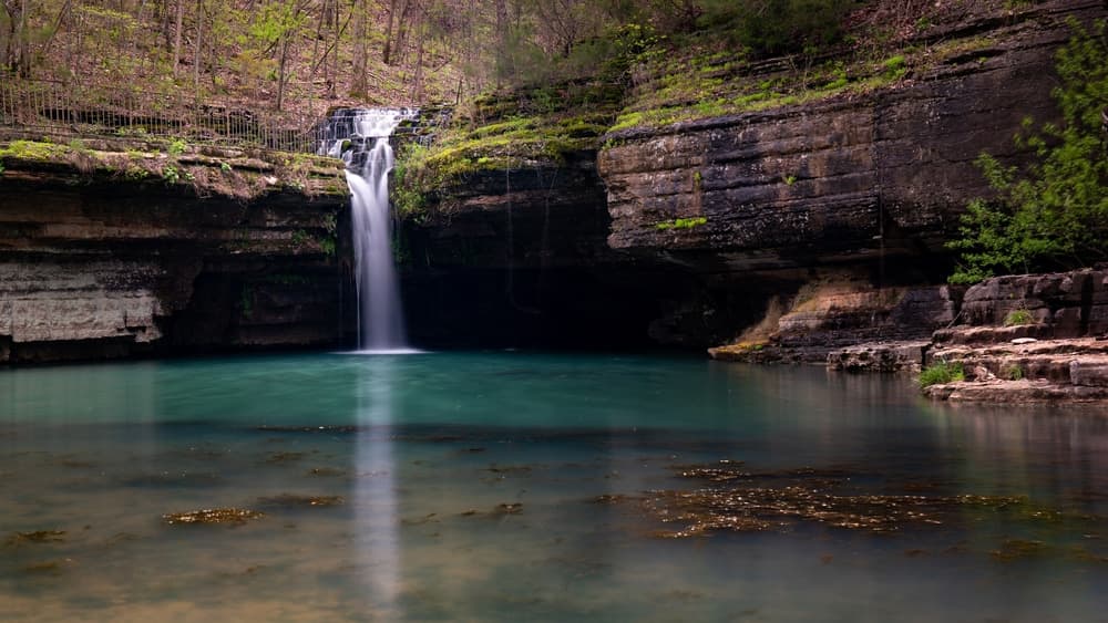 A serene waterfall flows into a calm turquoise pool surrounded by rocky cliffs and lush greenery.