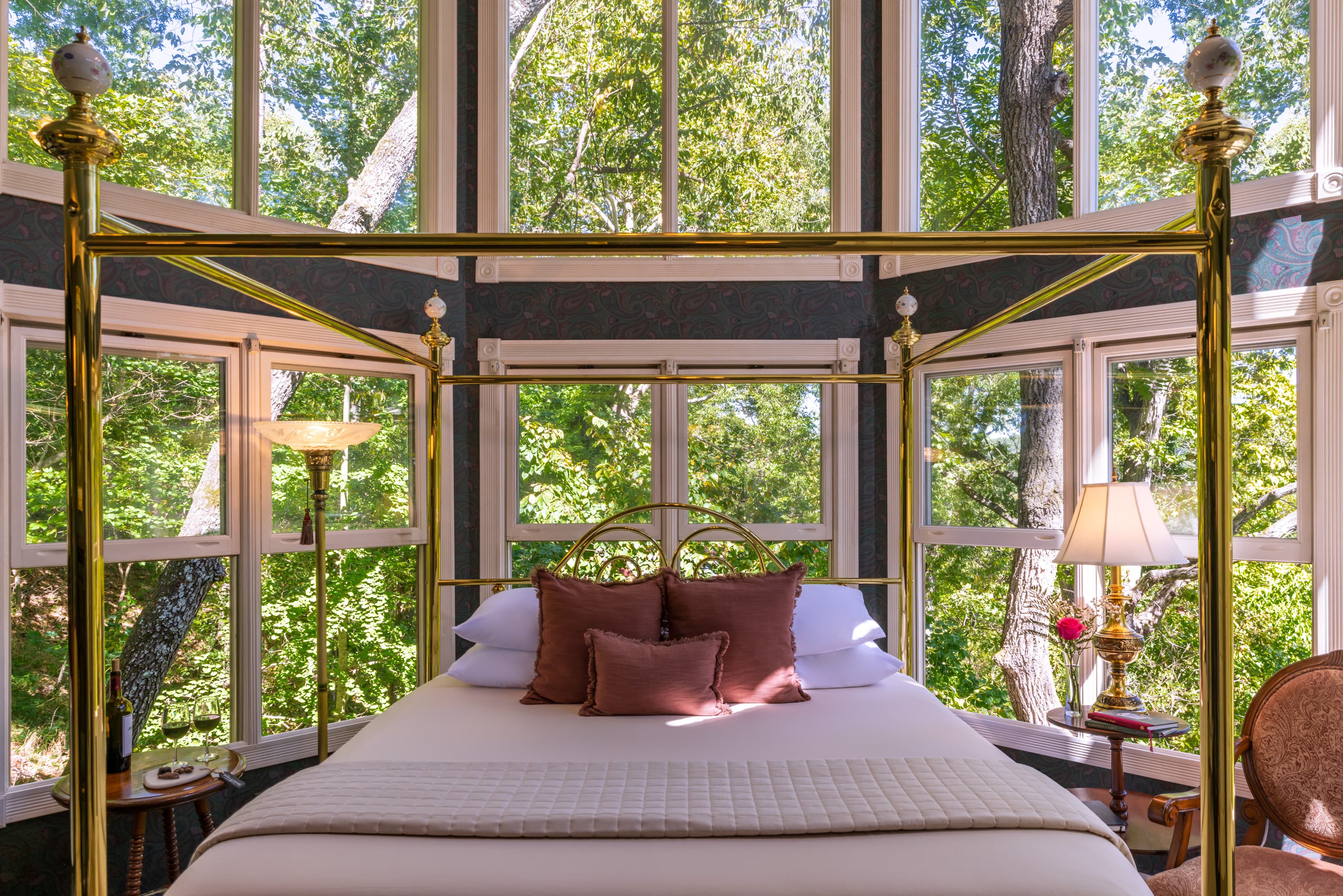 A cozy four-poster bed sits in a sunlit room surrounded by large windows overlooking lush greenery.