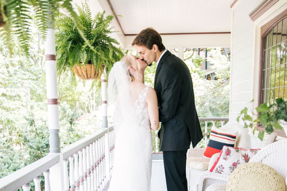 Beautiful bride and groom kissing on a white wrap-around porch with hanging fern.