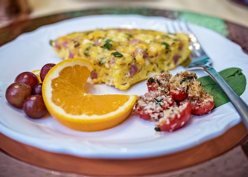 A plate featuring a slice of quiche, a slice of orange, a cluster of grapes, and a small dish of roasted tomatoes.