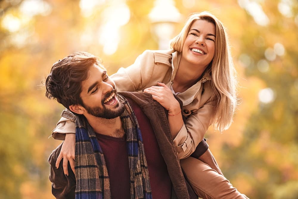 A man is laughing while giving a woman a piggyback ride in a sunlit autumn park.