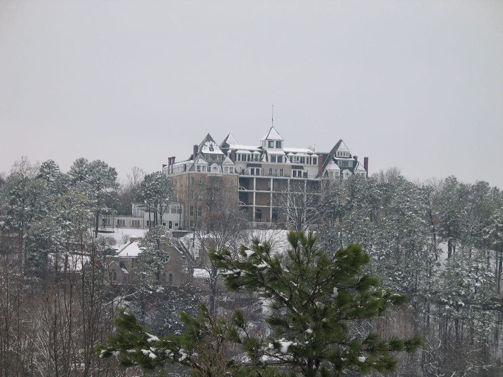 A large, historic building is covered in snow, surrounded by evergreen trees on a cloudy winter day.