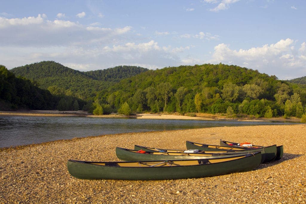 Four canoes resting on a pebbly riverbank with lush green hills in the background.