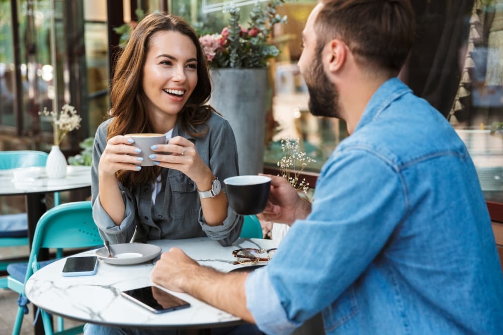 A woman and a man are smiling and enjoying coffee together at a café table.