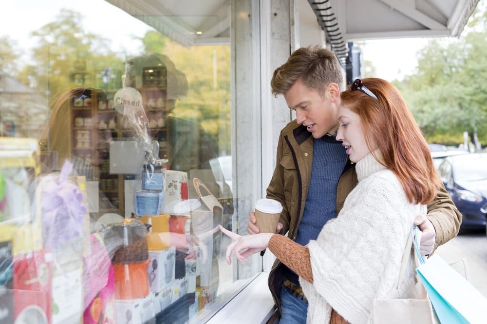 A couple looks at gifts through a store window, pointing and discussing items.