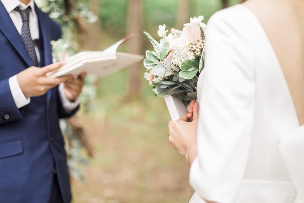 A couple stands in a lush outdoor setting, with the man reading from a book while the woman holds a bouquet.