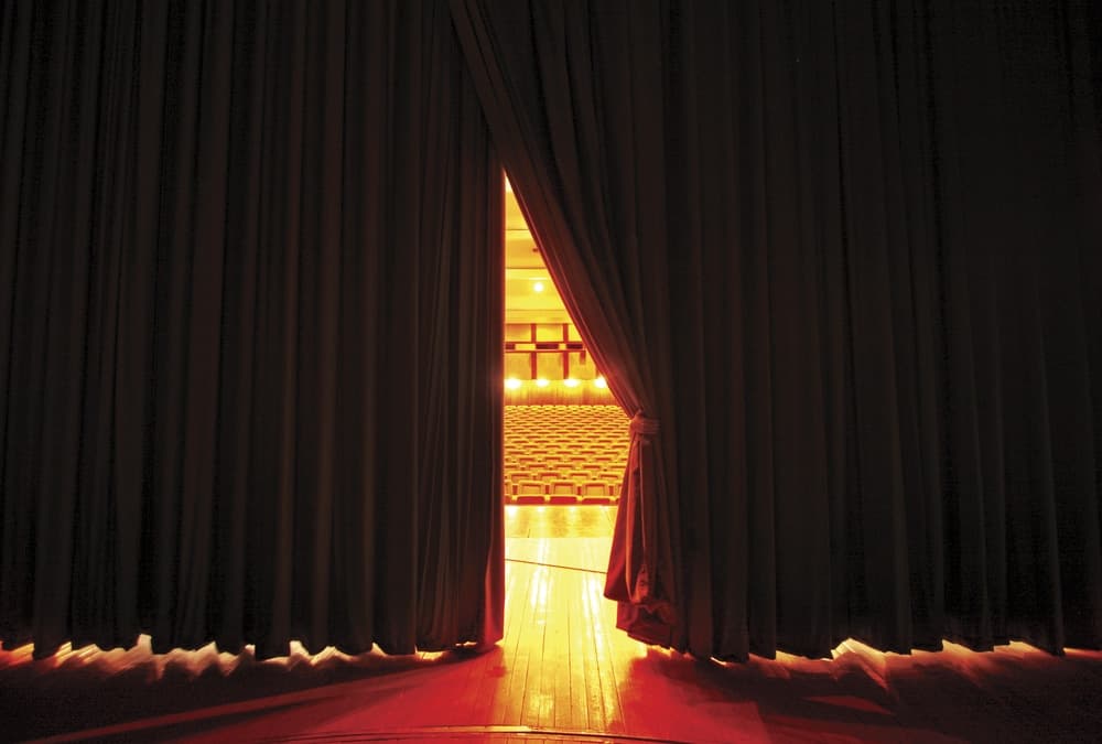 View from behind curtains looking out onto a brightly lit theater filled with empty seats.