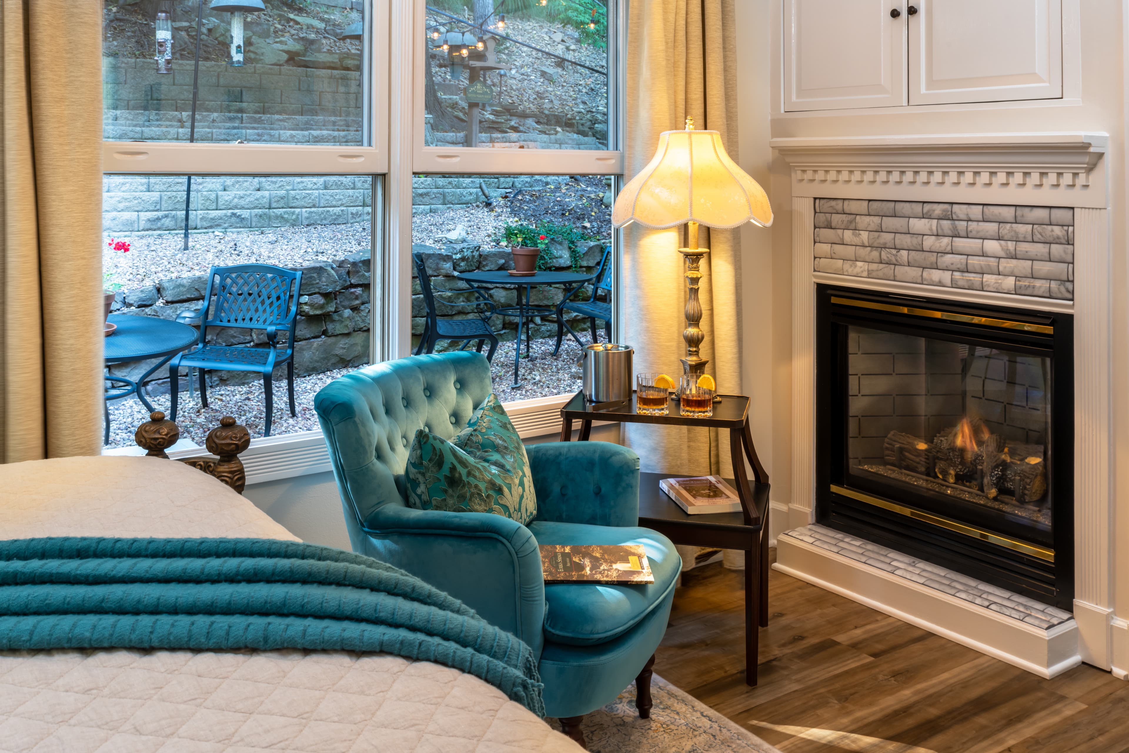 A cozy corner of a bedroom featuring an aqua chair, a lamp, and a fireplace, with a view of an outdoor seating area through large windows.