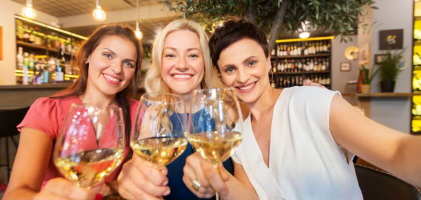 Three women smiling and toasting with glasses of wine in a vibrant bar setting.