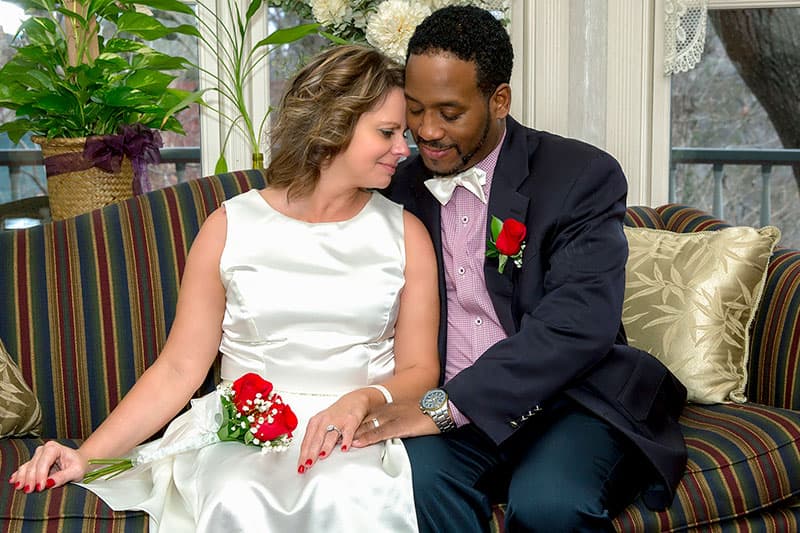 A couple in formal attire sits closely together on a couch, smiling and sharing an intimate moment.