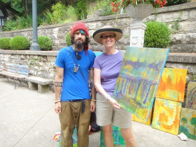 A man with a red beanie and beard stands next to a woman in a straw hat, both smiling while the woman holds a colorful painting.