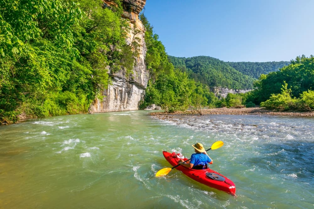 A person in a red kayak paddles on a river surrounded by lush green trees and rocky cliffs.