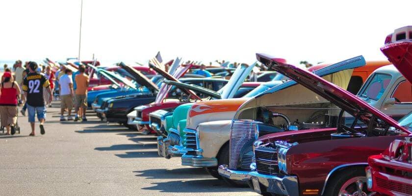 A row of classic cars with open hoods displayed at a car show, with spectators walking nearby.