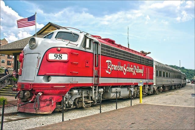 A vintage red train marked "Bronson Scenic Railway" is parked at a station with an American flag in the background.