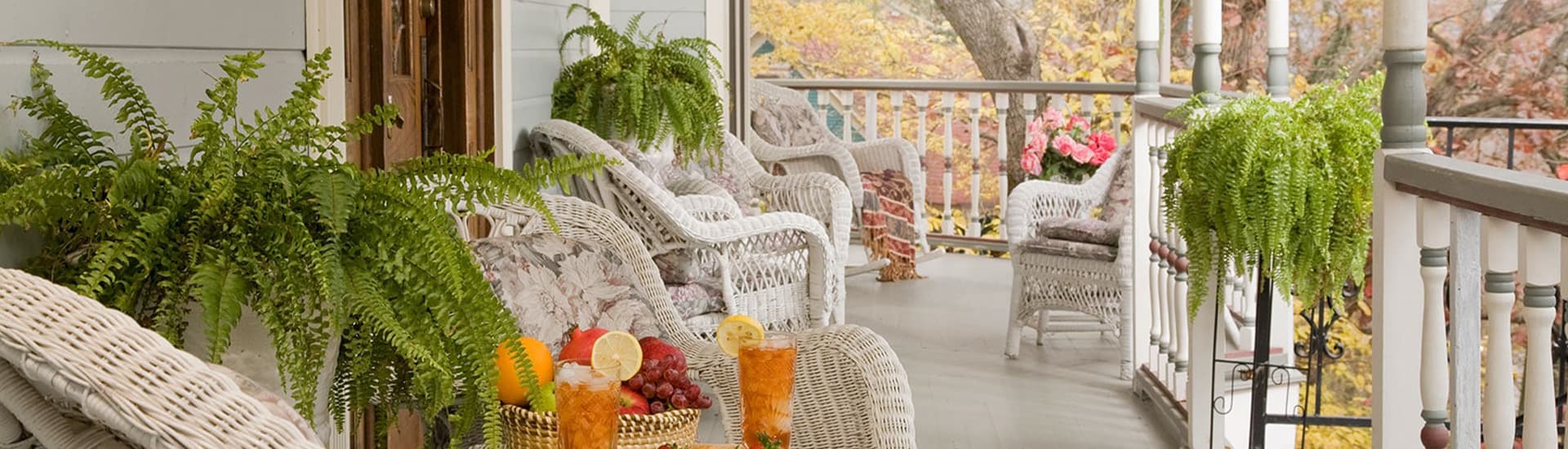 A spacious balcony with white wicker chairs, potted plants, and a basket of fruit and drinks. The balcony overlooks a colorful autumn landscape.