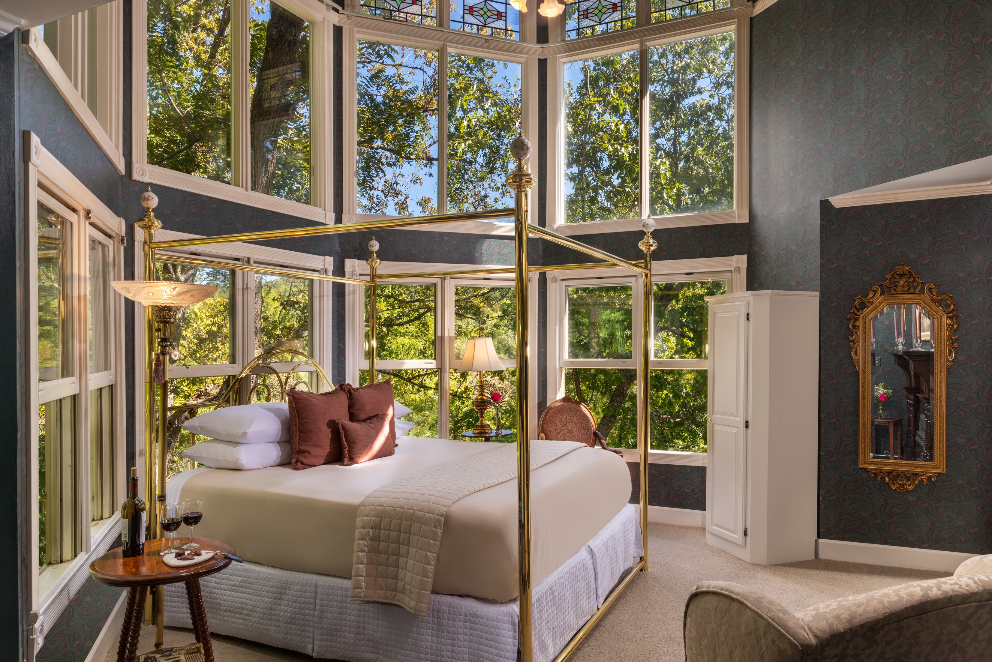 A bright bedroom featuring a four-poster bed, large windows with views of nature, and elegant furnishings.