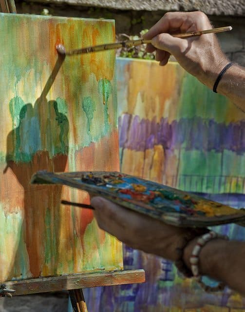 A person paints on a canvas while holding a palette of colorful paints.