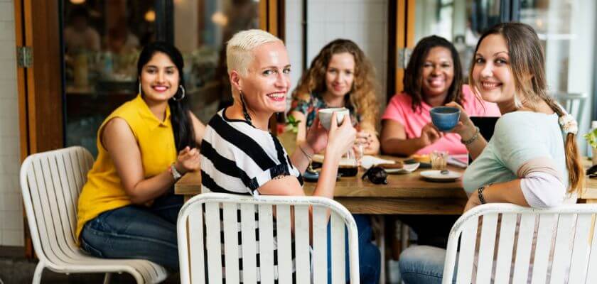 A group of five women smiling and enjoying drinks at a cafe table.