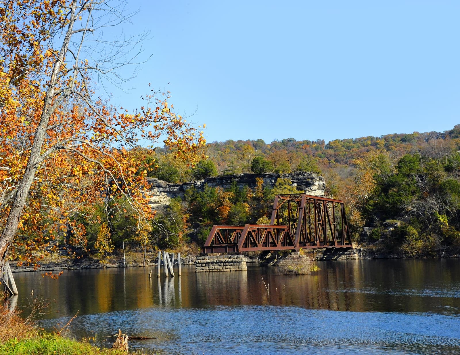 A rusted metal bridge spans a calm river surrounded by autumn foliage and rocky cliffs.