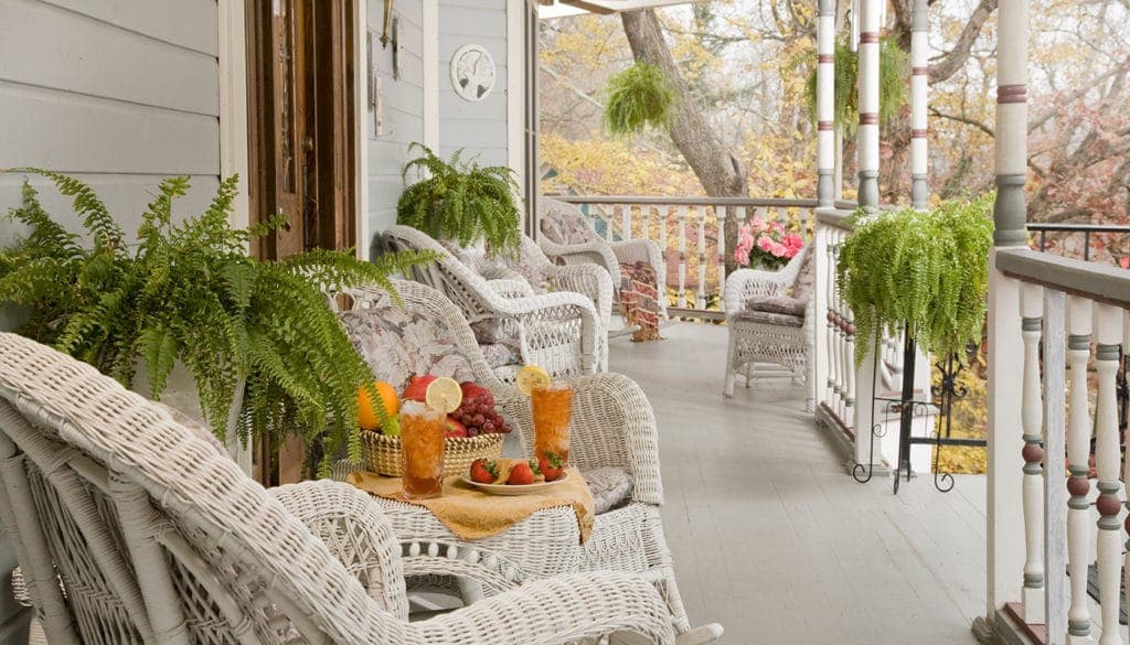 A cozy porch featuring white wicker chairs and a table with fresh fruit, surrounded by lush ferns and colorful plants.