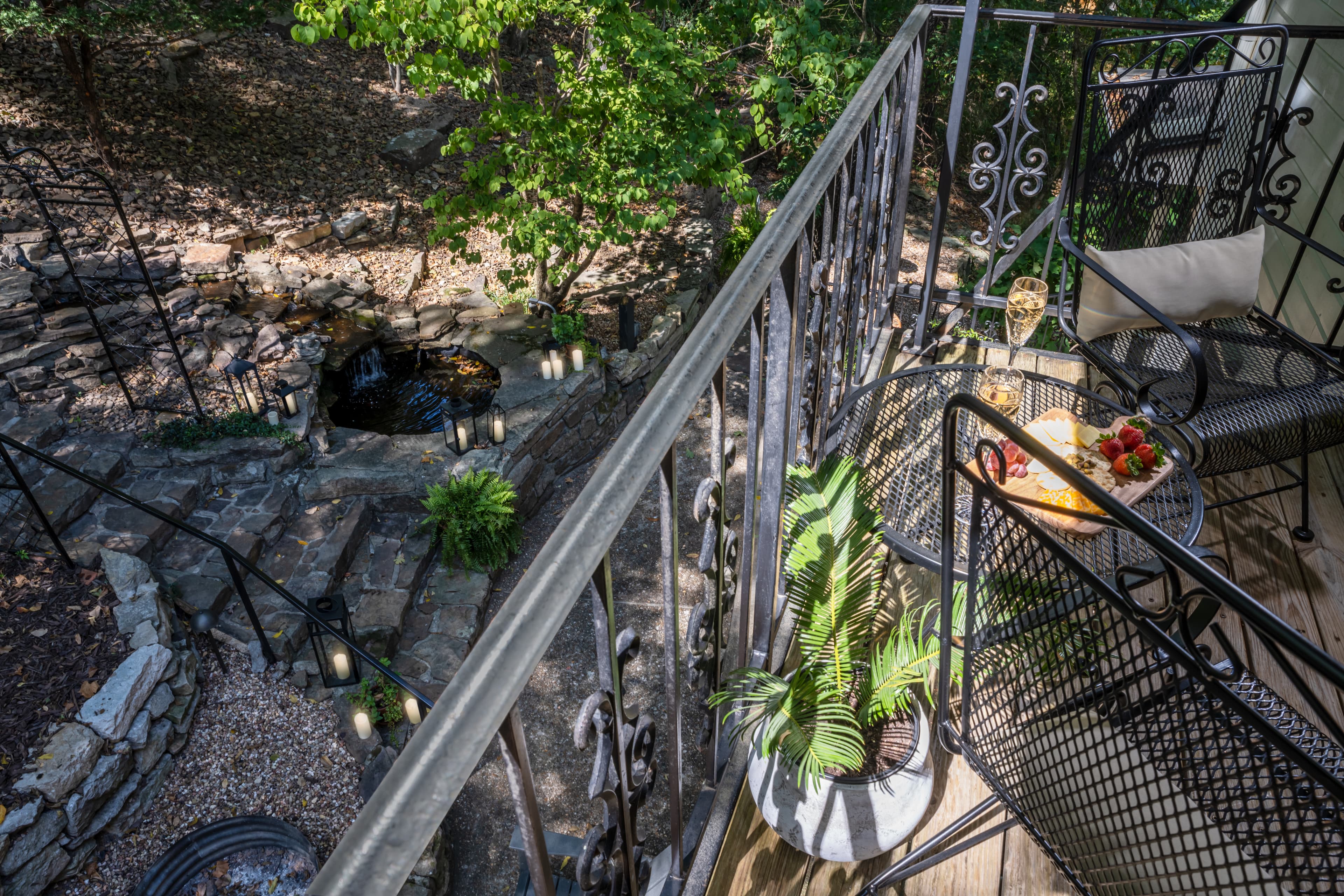 A cozy balcony overlooking a garden with a small pond, featuring two black chairs and a table with a plate of fruit.