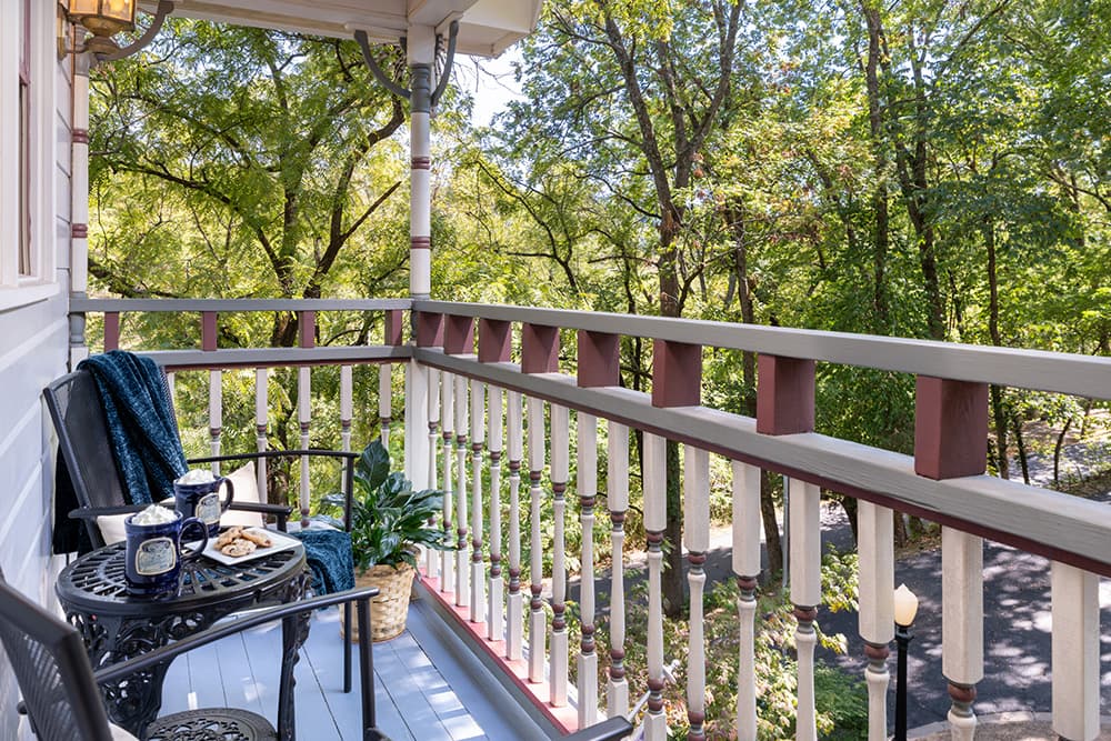 A cozy balcony with a view of a lush green forest. There is a small table with two chairs and a blanket on one of the chairs.