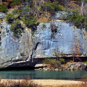 A rocky cliff adorned with sparse vegetation rises beside a tranquil river.
