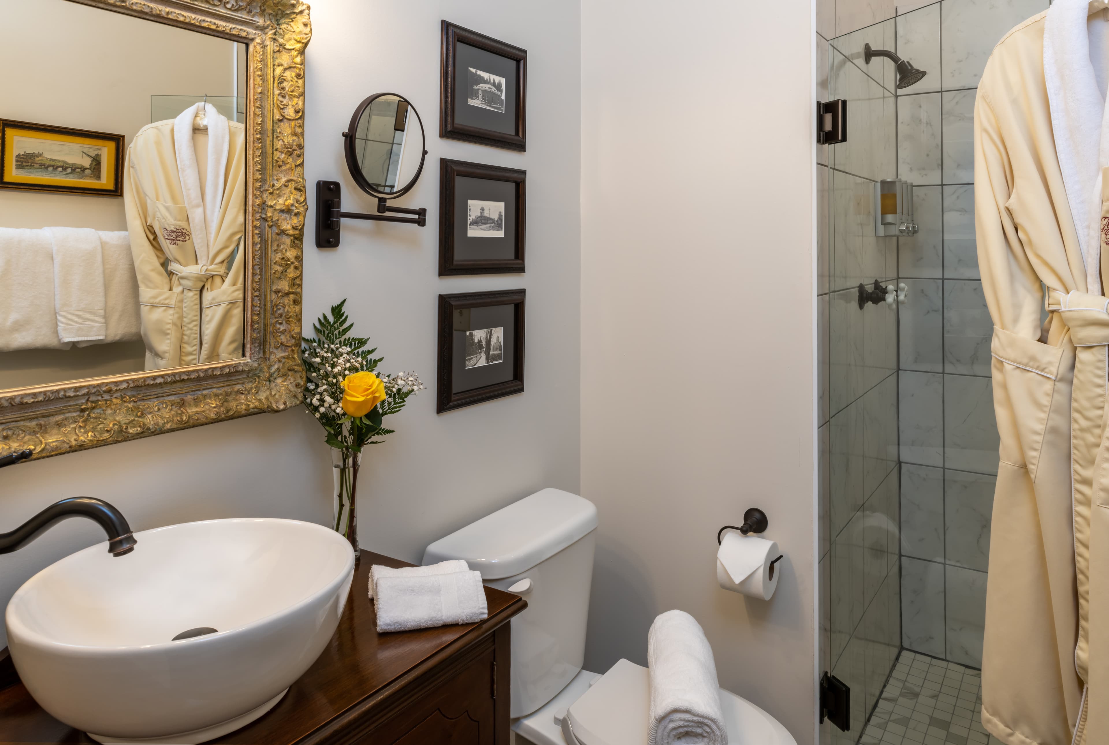 A neatly arranged bathroom featuring a decorative mirror, a flower vase, a plush robe hanging, and a spacious shower.