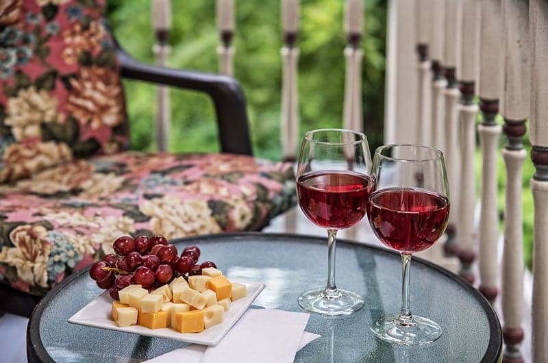 Two glasses of red wine sit beside a plate of cheese cubes and grapes on a glass table, with a floral-patterned chair in the background.