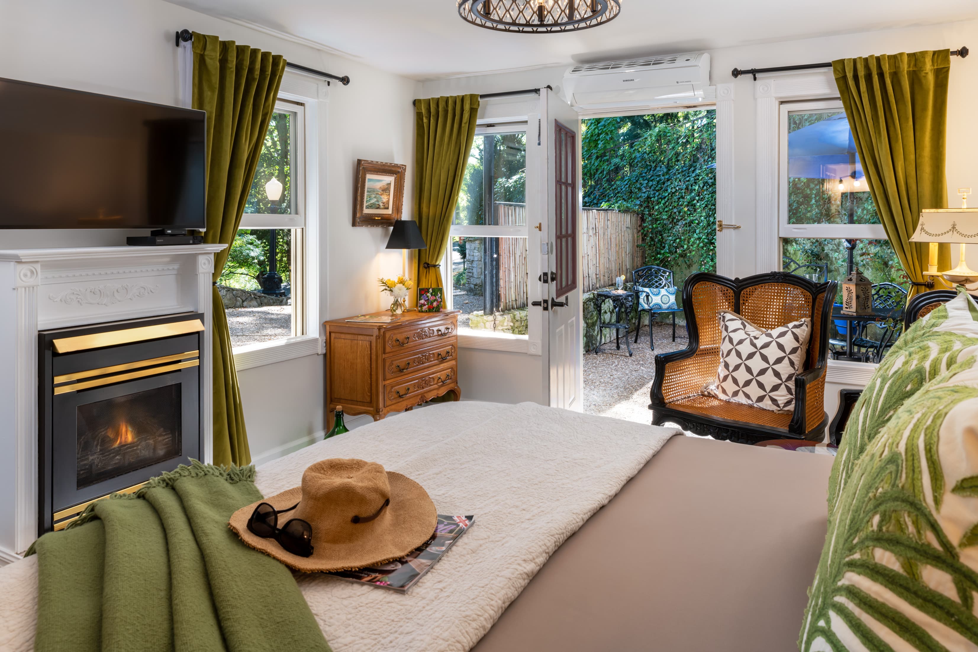 Cozy bedroom featuring a fireplace, large windows with green curtains, and outdoor seating visible through an open door.