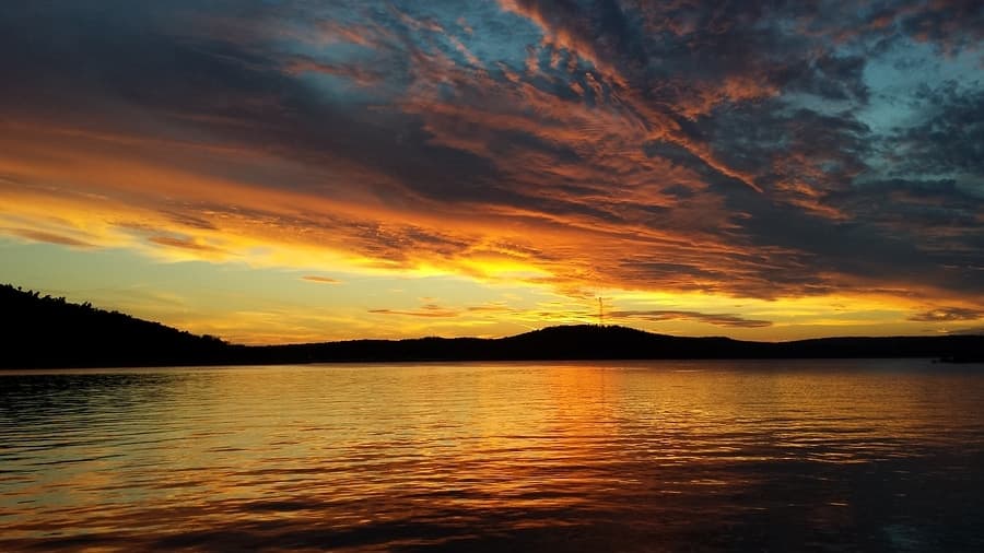 A colorful sunset reflects on calm water with silhouetted hills in the background.