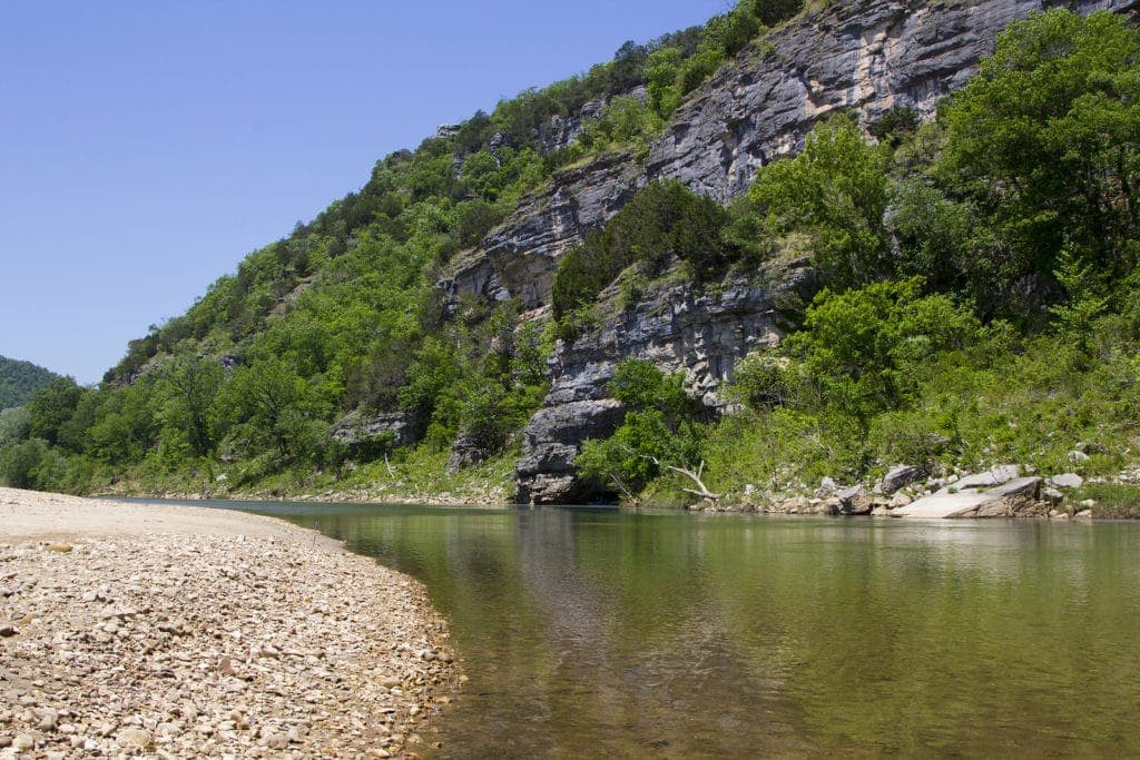 A serene river curves along a rocky shoreline, surrounded by lush greenery and steep cliffs under a clear blue sky.