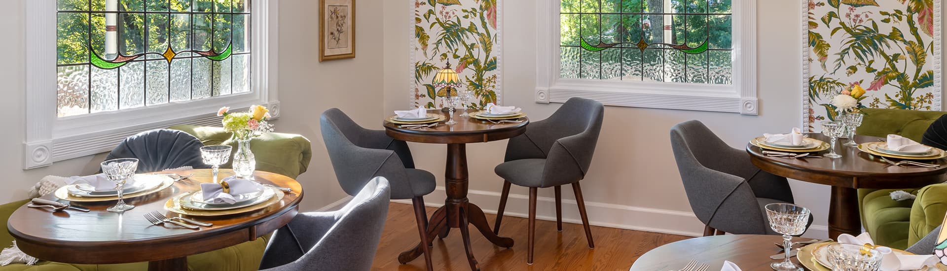 A cozy dining room with large windows, stained glass, and floral wallpaper.