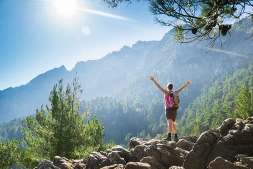 A person stands on a rocky ledge, arms outstretched, overlooking a mountainous landscape under a bright sun.