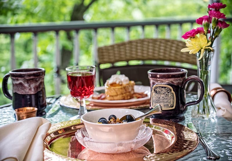 A table on a balcony overlooking a green garden. The table is set with two plates, mugs, a glass of wine, a bowl of fruit, and a vase of flowers.
