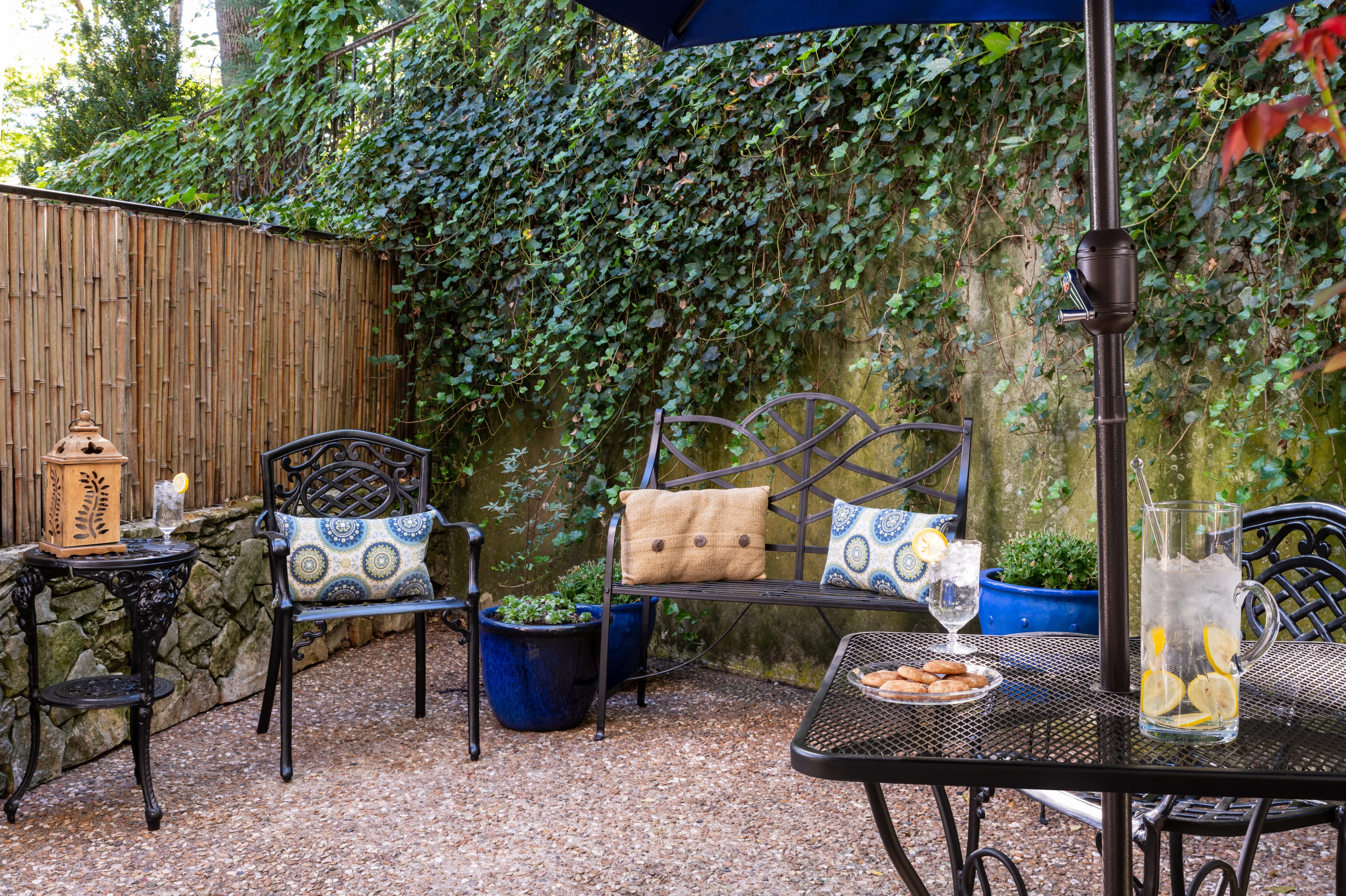 A cozy outdoor patio featuring two decorative chairs, a small black table with cookies, and lush greenery.