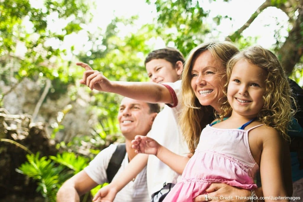 A family of four smiles and points at something off-camera in a lush outdoor setting.