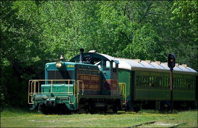 A green and blue vintage train is parked beside a signal in a lush, green landscape.