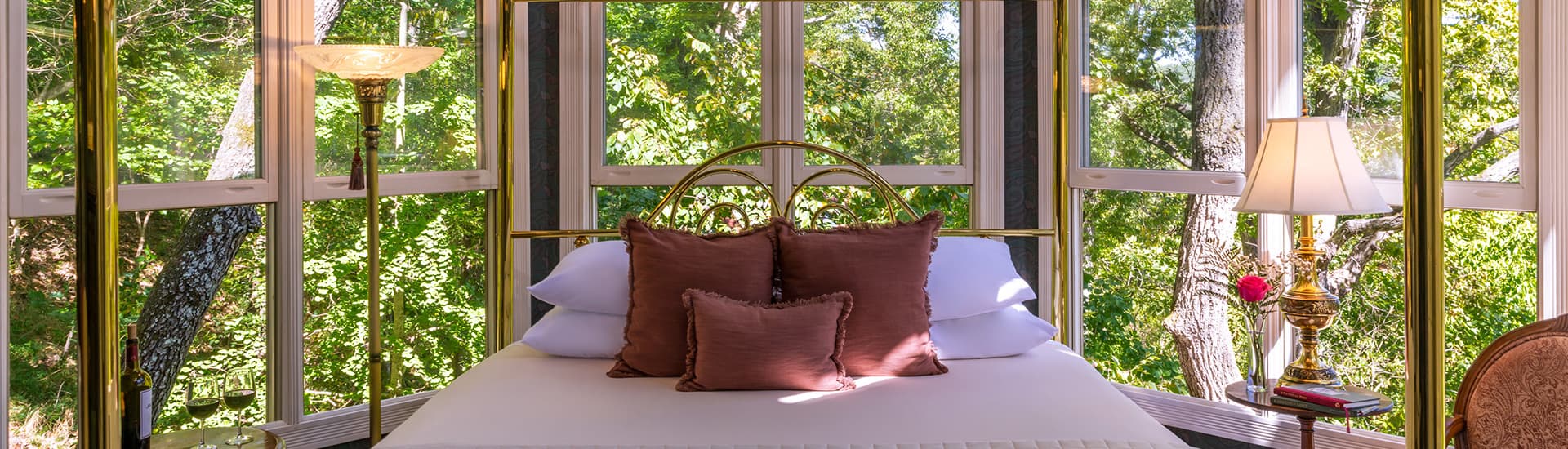 A luxurious bedroom with a canopy bed and ornate brass accents. The room is bathed in natural light from large windows overlooking a peaceful forest.