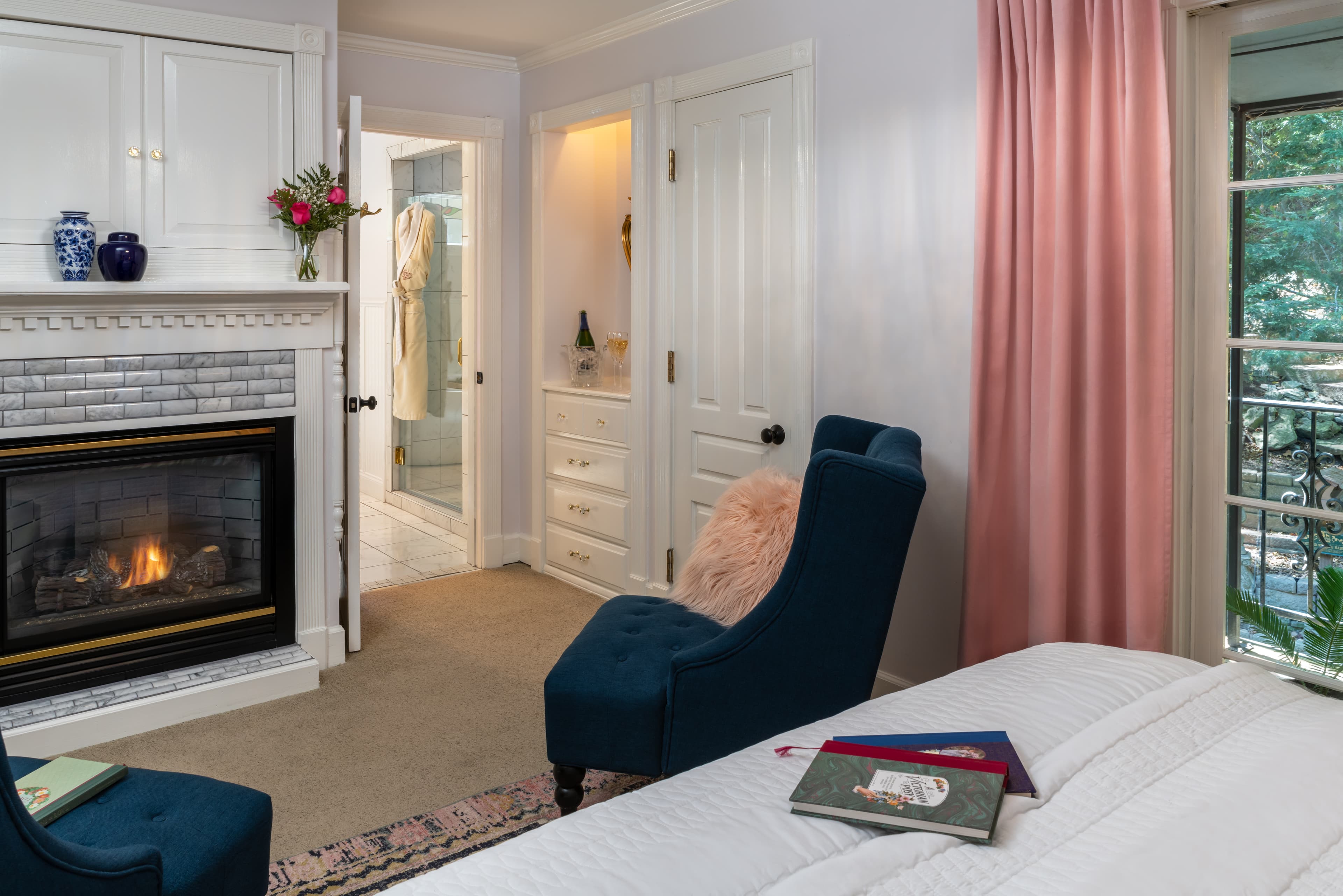 A cozy bedroom featuring a fireplace, blue chair, and soft pink curtains, with a bathroom visible in the background.