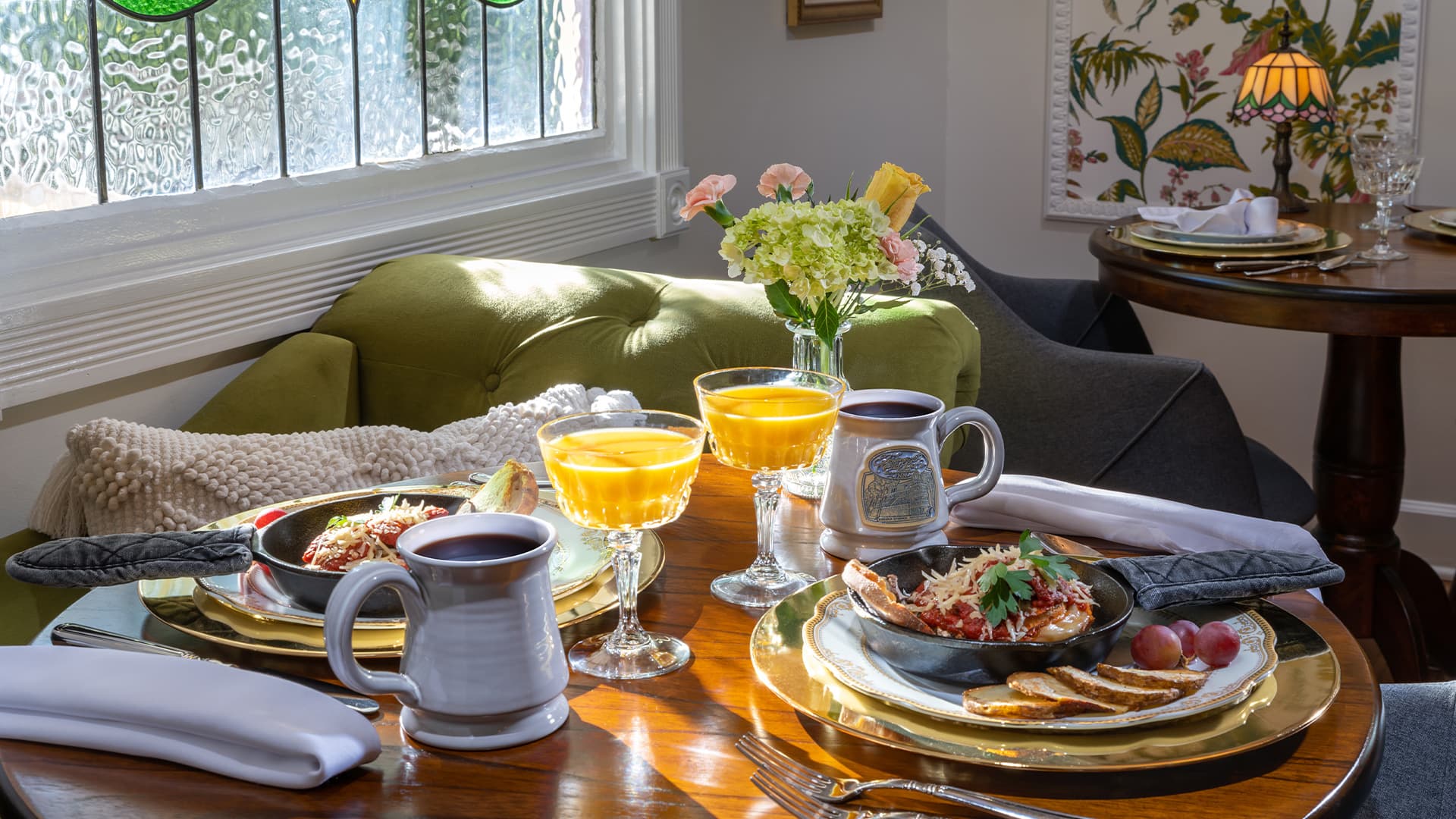 A round wooden table in a sunlit room with a stained glass window. The table is set for two with plates of food, mugs of coffee, glasses of juice, and a vase of flowers. A green sofa is visible in the background.