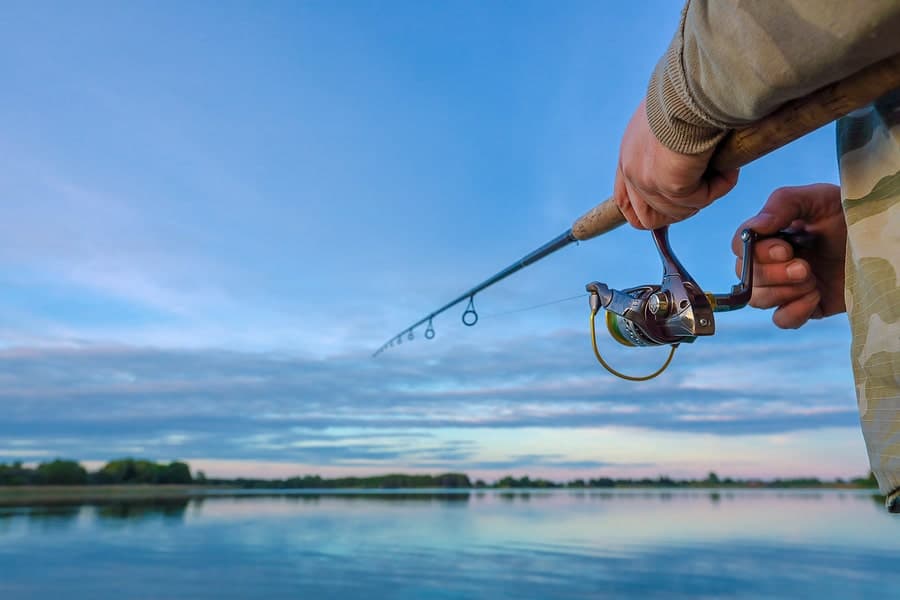 A person holds a fishing rod over a calm body of water under a blue sky.