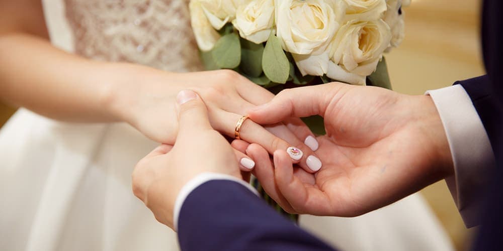A hand placing a wedding ring on another hand, with a bouquet of white roses in the background.
