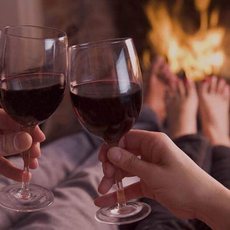 Two people holding glasses of red wine, sitting with their bare feet near a fireplace