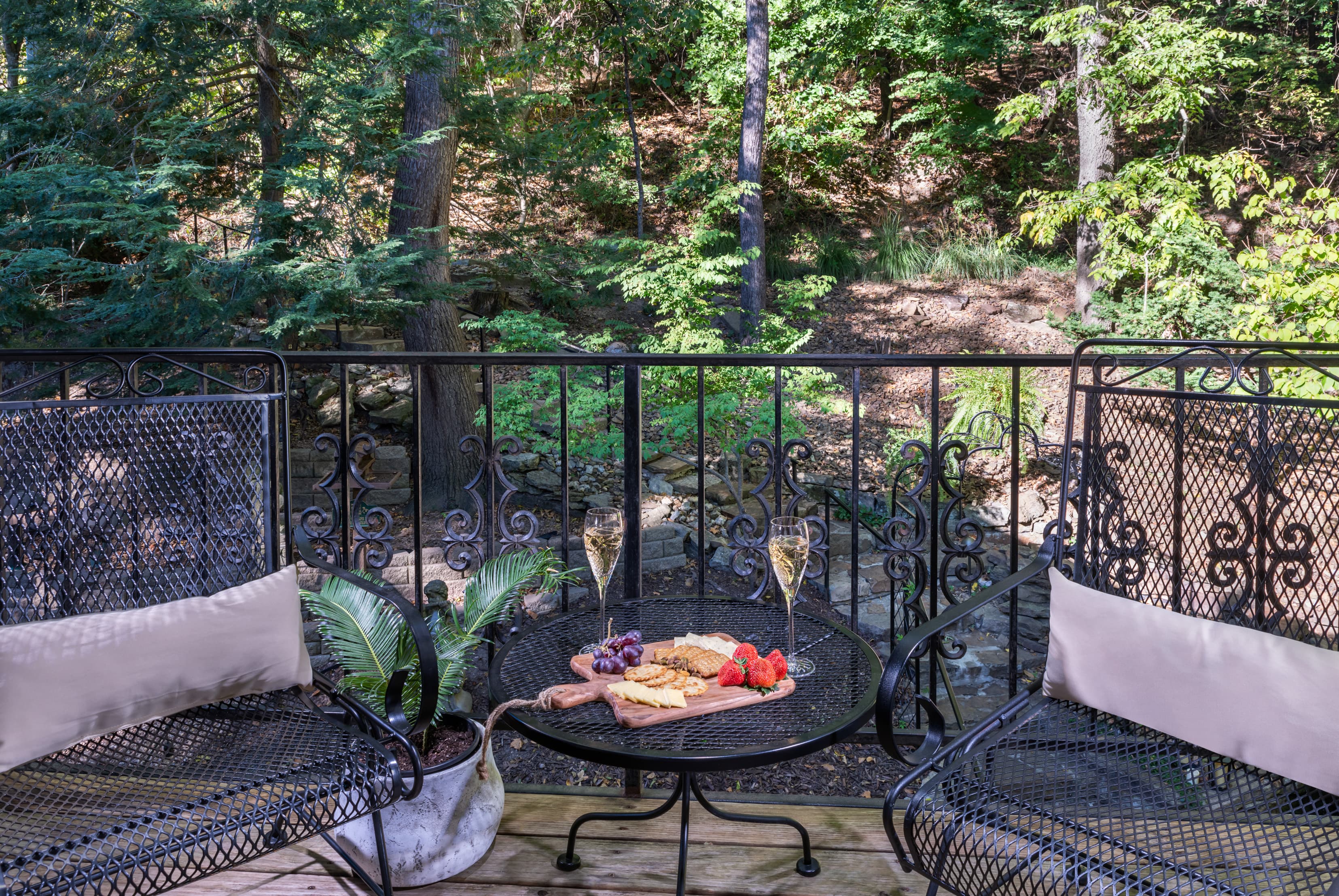 A cozy balcony features two chairs and a table set with snacks and two glasses of champagne, overlooking a lush green backyard.